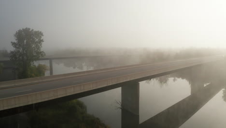 Heavy-fog-hangs-over-bridge-crossing-calm-river-with-early-morning-sunlight-shining-through