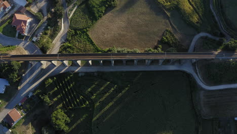 Crane-down-reveal-shot-of-old-arch-railway-bridge-surrounded-by-crop-fields,-casting-shadow-on-corn-field---Ponte-Seca,-Durrães,-Barcelos