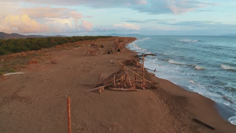 Aerial-drone-footage-of-pine-tree-forest-and-sandy-beach-seaside-in-the-iconic-Maremma-nature-park-in-Tuscany,-Italy,-with-a-dramatic-cloud-sky-at-sunset,-and-wooden-tipis-along-the-empty-beach