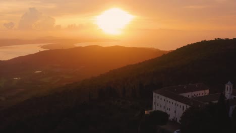 Aerial-drone-footage-of-the-sunrise-at-the-iconic-Monte-Argentario-lagoon,-facing-the-old-town-Orbetello-and-a-monastery-close-to-Maremma-nature-park-in-Tuscany,-Italy,-with-dramatic-clouds