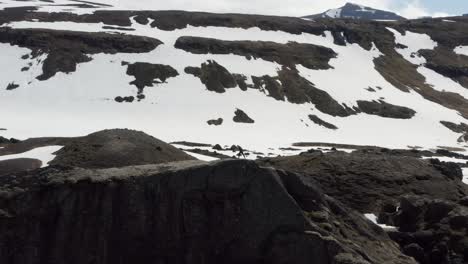 Mann-Sitzt-Auf-Einer-Bemerkenswerten-Grauen-Klippe-über-Wunderschönen-Türkisfarbenen-Gletscherbecken,-Steht-Auf-Und-Geht-Mit-Einem-Epischen-Schneebedeckten-Berg-Im-Hintergrund-Davon,-Drohnenantenne