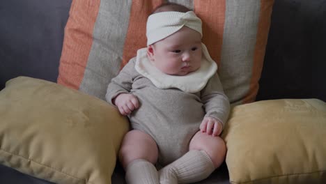 Adorable-baby-girl-sitting-alone-on-sofa-supported-with-cushions-wearing-a-bib-and-headband