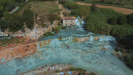 Aerial-drone-footage-of-the-iconic-Saturnia-Cascate-del-Mulino-thermal-hot-springs-with-its-blue-warm-water-in-the-idyllic-landscape-of-Tuscany,-Italy-with-hills,-fields,-people-and-olive-trees-around