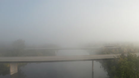 Drone-backing-away-from-highway-bridges-crossing-peaceful-river-ascends-into-fog-hanging-over-the-landscape