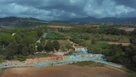 Aerial-drone-footage-of-the-iconic-Saturnia-Cascate-del-Mulino-thermal-hot-springs-with-its-blue-warm-water-in-the-idyllic-and-romantic-landscape-of-Tuscany,-Italy-with-fields-and-olive-trees-around