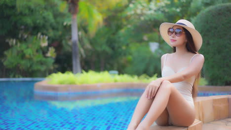 slow-motion-of-young-Asian-woman-sitting-near-the-pool-in-tropic-resort-touching-the-frame-of-sunglasses,-wellness-concept
