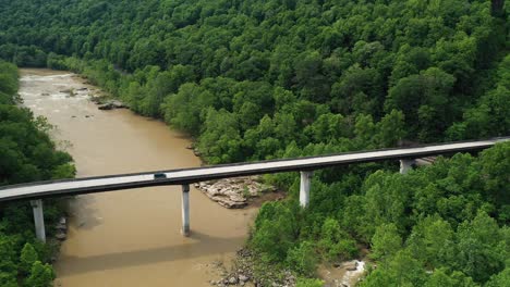 Dark-Van-Passing-Bridge-Above-Muddy-River,-Aerial-View,-Lush-Green-Forest-and-Roadway-in-West-Virginia-USA