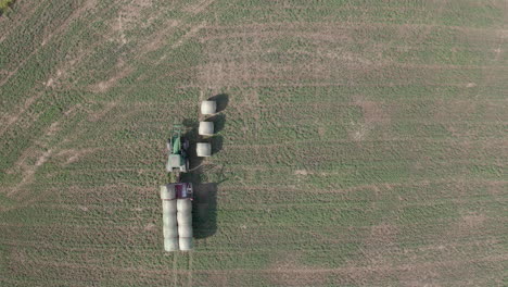 View-directly-above-green-industrial-tractor-machine-loading,-collecting,-gathering-and-hauling-hay-bales-in-rural-flat-plains-farmland-and-countryside,-Saskatchewan,-Canada,-overhead-drone-static