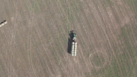 Agricultural-farmer-driving-green-industrial-tractor-machine-hauling-hay-bales-in-rural-countryside-flat-plains-farmland,-Saskatchewan,-Canada,-directly-above-circle-aerial-descend