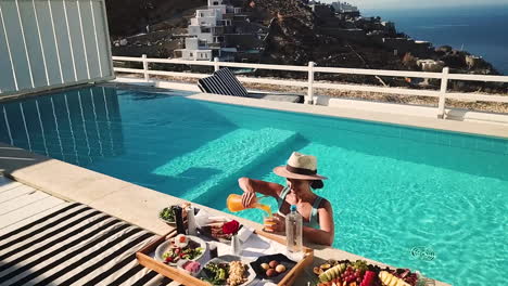 Aerial-View-of-Happy-Girl-in-Private-Pool-Pouring-Orange-Juice-on-Breakfast-Tray-on-Sunny-Day-in-Luxury-Mykonos-Villa,-Slow-Motion