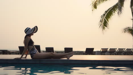Zeitlupe-Auf-Der-Silhouette-Einer-Asiatischen-Modellfrau,-Die-Bei-Sonnenuntergang-Im-Thailändischen-Resort-Am-Rand-Des-Swimmingpools-Sitzt,-Seitenansicht
