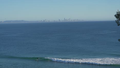 Surfer-Surfing-On-The-Calm-Blue-Sea-In-Currumbin---Distant-View-Of-Gold-Coast-City-Skyline-In-The-Background---Surfing-In-Queensland,-Australia---wide-shot