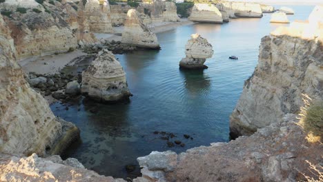 Slowly-Tilting-up-shot,-Scenic-view-of-Rock-Tunnels-beach-in-Algarve,-Portugal,-people-walking-on-the-beach-and-boat-floating-near-the-rock-formation-on-the-sea