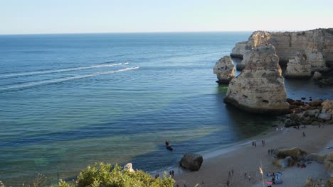 Einspielung,-Schnellbootrennen-Auf-Dem-Meer-Der-Algarve,-Portugal,-Malerischer-Blick-Auf-Die-Menschen-Am-Felstunnelstrand-Im-Hintergrund