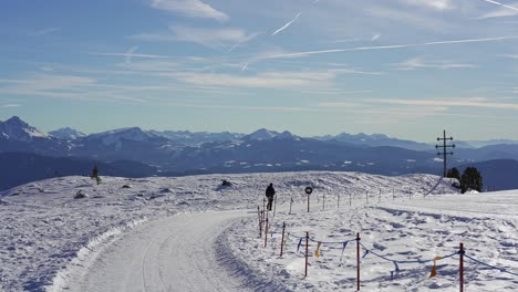 Caminante-Camina-Solo-A-Lo-Largo-De-Un-Camino-De-Nieve-En-La-Cima-De-Una-Montaña-Cubierta-De-Nieve-Desierta-Con-Picos-De-Montaña-épicos-En-El-Fondo