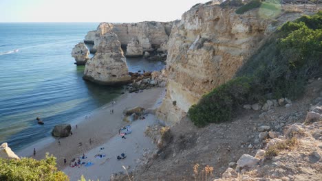 Tilting-up-shot,-Scenic-view-of-People-bathing-on-the-coastline-of-Rock-tunnel-beach-in-Algarve,-Portugal,-waves-hitting-the-rock-formation-in-the-background