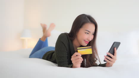 Schöne-Thailändische-Frau,-Die-Auf-Einem-Kingsize-Luxusbett-Liegt-Und-Die-Kreditkartennummer-In-Ihr-Mobiltelefon-Eingibt,-Um-Online-Zu-Bezahlen