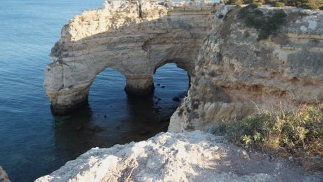 Tilting-up-shot,-Scenic-view-of-Rock-tunnel-cliff-in-Algarve,-Portugal,-People-standing-on-the-top-of-the-cliff-on-the-bright-sunny-day