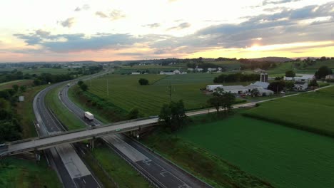 Cars-and-trucks,-on-highway-under-dramatic-sunrise,-sunset-among-green-rural-American-farmland-during-summer,-rising-aerial-of-traffic