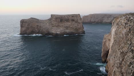 Einspielung,-Malerischer-Blick-Auf-Eine-Welle,-Die-Die-Felsformation-In-Der-Nähe-Der-Küstenlinie-Der-Algarve,-Portugal,-Trifft,-Bewölkter,-Nebliger-Himmel-Im-Hintergrund