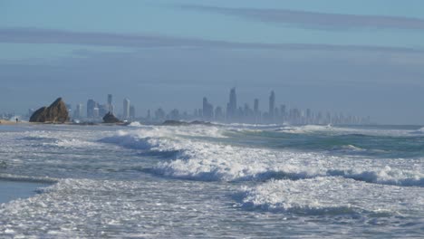 Surfing-Waves-Rolling-Over-The-Sea-In-Currumbin-Beach-On-A-Summer-Morning---Gold-Coast-Skyline-In-The-Misty-Background---wide-shot