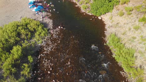 Flying-over-a-swimming-hole-up-to-a-bridge-in-the-American-River-in-Auburn,-California---surrounded-by-green-trees,-mountains-and-filled-with-people-swimming-and-enjoying-their-summer-weekend