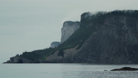 Gorgeous-Foggy-Morning-Overlooking-an-Epic-Costal,-Tree-Layered-Cliffside-at-Forillon-National-Park,-Canada-with-Calm-Gulf-of-St-Lawerence-Waters-in-the-Foreground