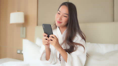 A-medium-close-of-a-pretty-young-woman-sitting-on-the-bed-in-her-luxury-hotel-suite-while-wearing-a-resort-bathrobe,-enjoys-communicating-through-her-smartphone
