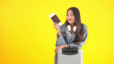Smiling-asian-woman-holding-passport-and-air-ticket-near-suitcase-isolated-on-yellow-background