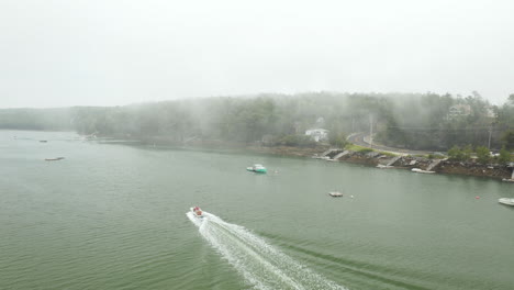 Fly-Over-Drone-Aerial-Footage-over-coast-of-Harpswell,-Cumberland-County,-Maine,-showing-speed-boat-crossing-water-parallel-to-coastline-on-foggy-day