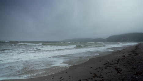 An-Einem-Deprimierend-Kalten-Winterlichen-Morgen,-Perce-Quebec,-Rollen-Wellen-Herein-Und-Erzeugen-Meeresschaum-An-Einem-Leeren-Strand-Mit-Nebelbedeckten-Meeresklippen-Im-Hintergrund
