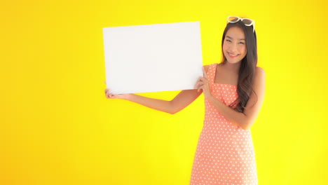 Young-Asian-woman-in-summer-dress-with-sunglasses-on-head-holds-up-plain-white-sign-board-and-smiles