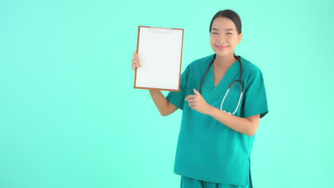Young-Asian-model-in-medical-scrubs-with-stethoscope-holds-up-clipboard-with-blank-white-page,-smiles-and-gives-thumbs-up