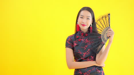 Mujer-Asiática-Vestida-Con-Traje-Tradicional-Chino-Sosteniendo-Un-Abanico-De-Encaje-Negro,-Aislada-De-Fondo-Amarillo