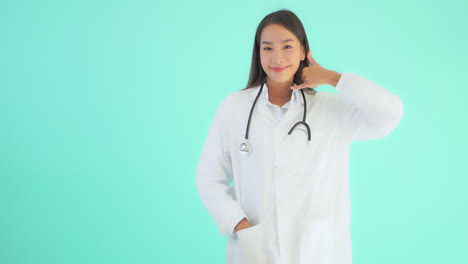 Smiling-Asian-model-in-white-lab-coat-with-stethoscope-uses-hand-to-signal-making-a-call-and-points