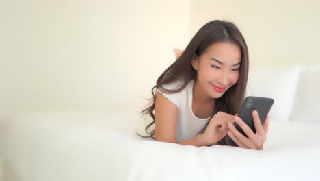 Simplistic-white-concept,-woman-typing-on-her-mobile-phone-while-lying-on-the-cozy-king-size-bed-facing-the-camera-smiling,-camera-slide-up