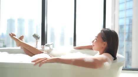 Young-Asian-Woman-taking-a-hot-bath-relaxing-in-a-bathtub-filled-with-foam-on-the-high-floor-of-a-luxury-hotel-with-ceiling-to-floor-windows