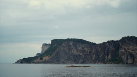 Serene-Calm-Sea-Waters-sit-in-front-of-an-Epic-Huge-Flora-Covered-Ocean-Cliffside-on-an-Overcast-Wintery-Day-at-Forillon-National-Park,-Canada