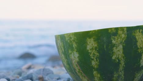 Die-Hälfte-Der-Reifen-Wassermelone-Auf-Den-Kieselsteinen-Am-Strand-Mit-Plätschernden-Wellen-Im-Hintergrund-In-Griechenland