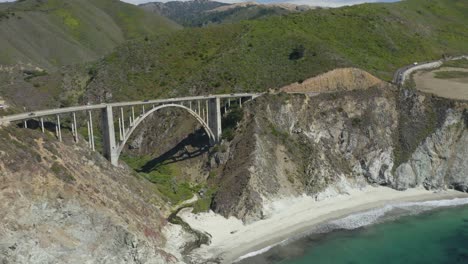 California's-Famous-Bixby-Creek-Bridge,-Waves-Crashing-on-Beach-Below,-Mountains-in-Background