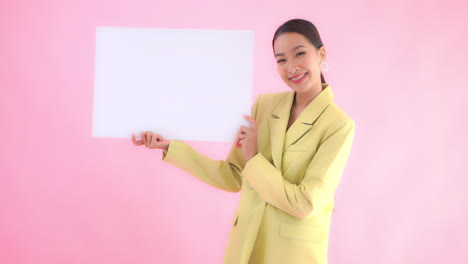 Asiatische-Geschäftsfrau,-Die-Ein-Leeres-Blatt-Des-Papierplakatbanners-Mit-Einem-Glücklichen-Gesichtsausdruck-Auf-Rosa-Studiohintergrund-Hält