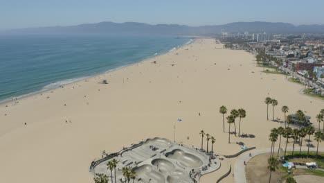 Drone-Flies-Over-Venice-Beach-with-Skatepark-and-Palm-Trees-Below-on-Summer-Day
