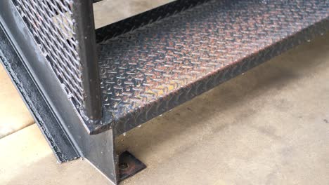 Rain-dripping-slowly-off-of-textured-industrial-metal-cross-plate-steel-staircase-outside-onto-cement-ground-in-slow-motion