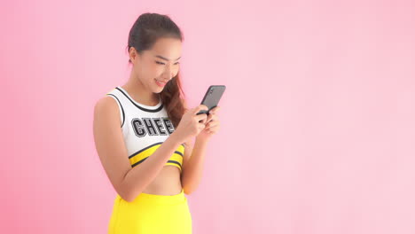 Attractive-Asian-model-in-cheerleader-costume-using-phone-with-plain-pink-background-and-copy-space-on-right