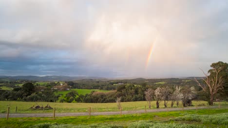 Eine-Wunderschöne-Zeitrafferaufnahme-Von-Grünen-Sanften-Hügeln-In-Australien-Mit-Dicken-Wolken,-Die-Im-Abendlicht-Leuchten,-Und-Einem-Regenbogen,-Der-Am-Himmel-Erscheint