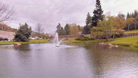 Canada-goose-flies-away-by-camera-as-drone-flies-forward-through-a-pond-and-water-fountain-surrounded-by-green-trees-and-grass-during-an-overcast-cloudy-day-by-The-Lake-of-Pines-in-Auburn,-California