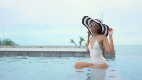 Happy,-attractive-Asian-woman-with-large-hat-and-sunglasses-sitting-on-ledge-inside-infinity-swimming-pool-of-luxury-tropical-island-resort-with-ocean-in-background