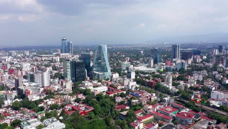 Aerial-backward-flight-above-downtown-Mexico-city-center-skyscrapers,-buildings-and-Plaza-Manacar-mall-with-mountain-range-in-background-on-cloudy-sky-day,-overhead-drone-pull-back