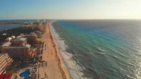 Tropical-sea-waves-crashing-on-white-sandy-shore-with-a-blue-sky,-golden-sun-rays-shine-on-turquoise-sea-next-to-Hotel-Zone-resorts---Laguna-Nichupté-in-Cancun,-Mexico---Drone-still-4k