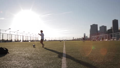 Wide-Shot-of-Girl-Silhouetted-Juggling-Soccer-Ball-with-Sun-and-Cityscape